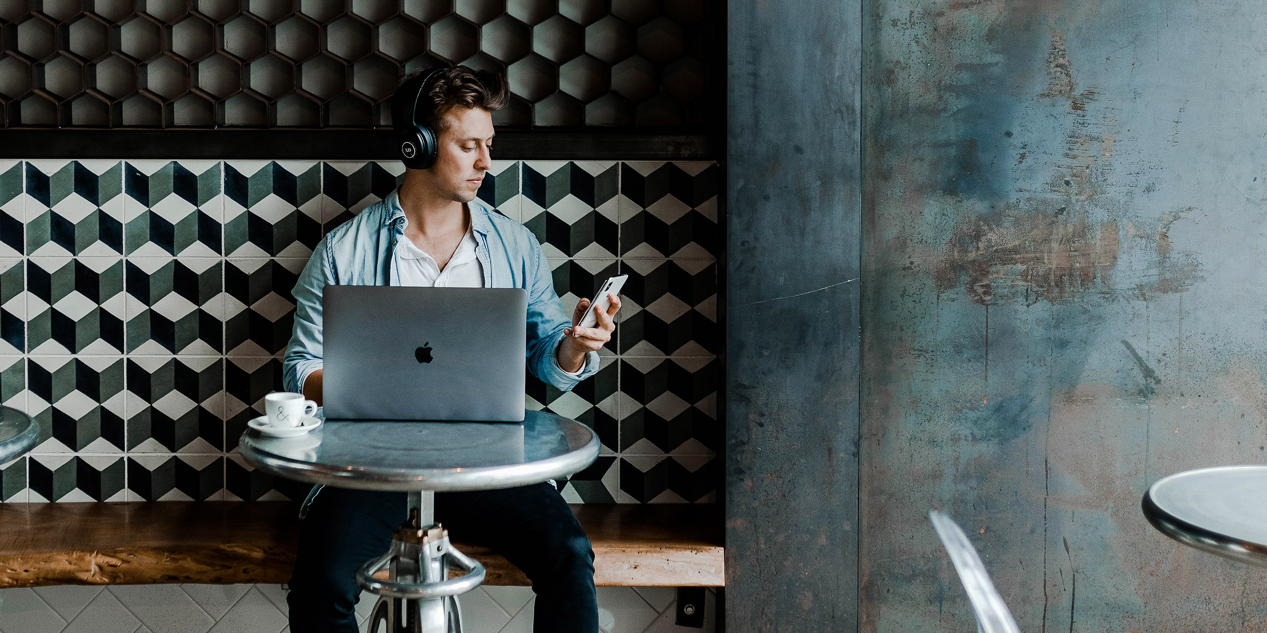 Image of young man working on laptop in a cafe while wearing wireless headphones and glancing at his mobile phone